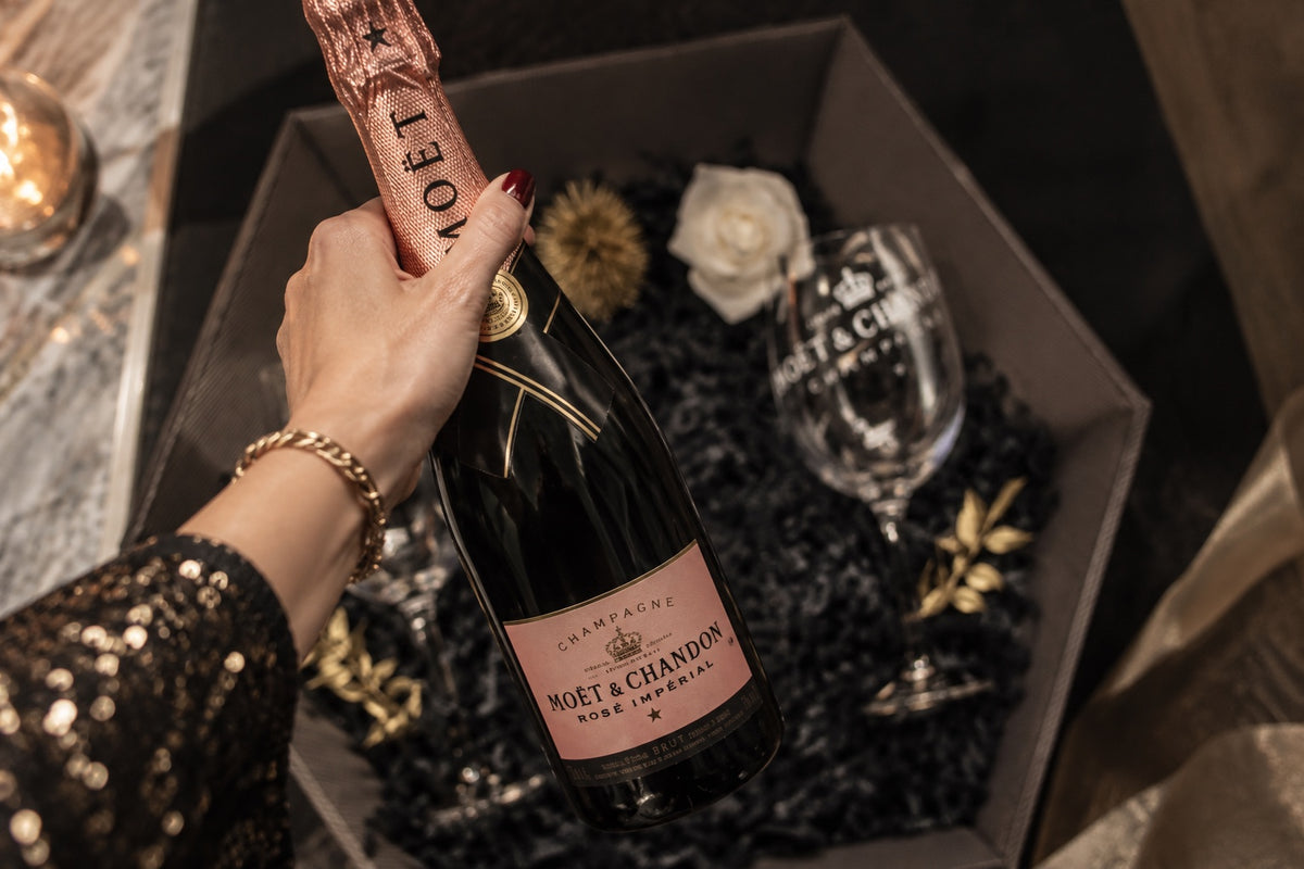 Hand hält eine Flasche Moët & Chandon Rosé Impérial über einer schwarzen Geschenkbox mit Glas und weißer Rose
